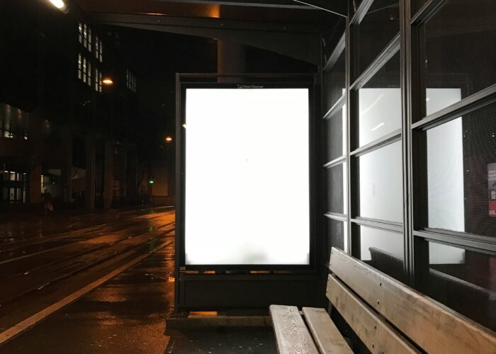 A glowing blank advertising billboard at a wet bus stop at night